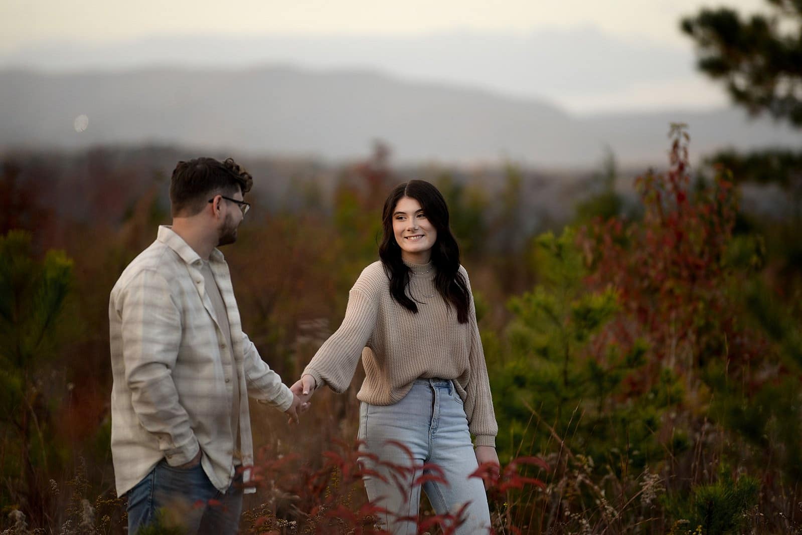 A North Georgia couple holding hands in tall autumn grass with mountain bokeh and fall foliage behind them — engagement photo outfit example